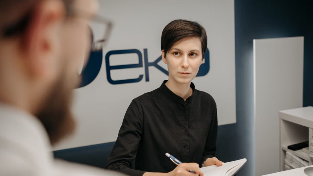 A receptionist at a clinic desk writing notes during a consultation.